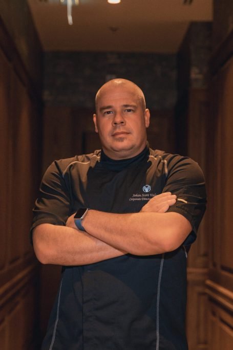 Bald chef standing confidently indoors, arms crossed, in a stylish hallway.
